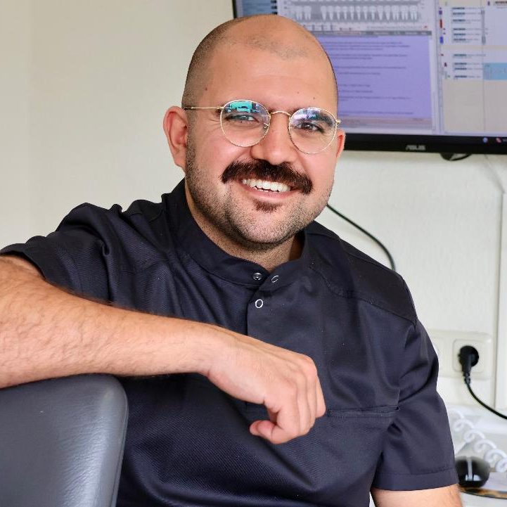 Lächelnder Mann mit Brille und Schnurrbart, sitzt in einem modernen Zahnarztbehandlungsraum.