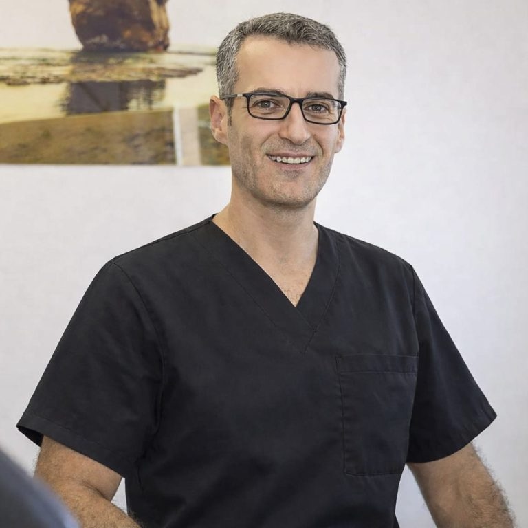 Lächelnder Mann mit Brille und Schnurrbart, sitzt in einem modernen Zahnarztbehandlungsraum.